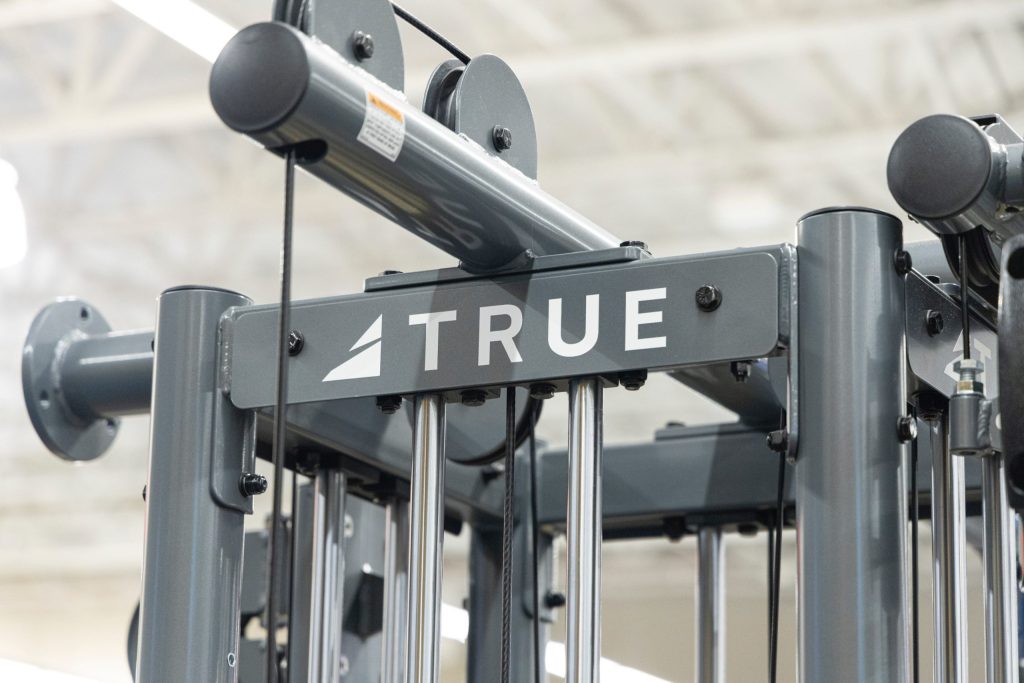 Close-up of a gray gym machine with metal bars and pulleys, featuring the brand name “TRUE” on a label in the center. The background is blurred, showing a bright, indoor gym environment.
