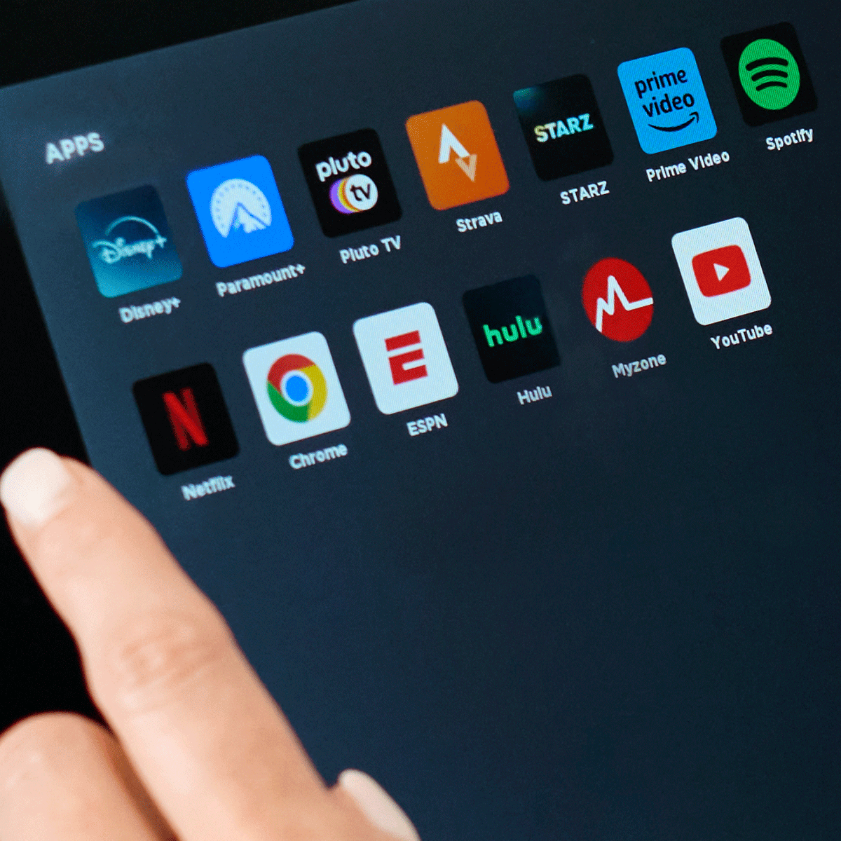 A person’s hand points at a tablet screen displaying various app icons, including Disney+, Netflix, Chrome, YouTube, Hulu, Spotify, ESPN, Prime Video, Paramount+, Pluto TV, Strava, STARZ, and Myzone.