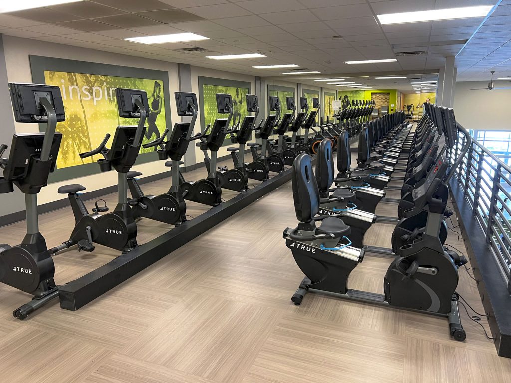 Rows of stationary bikes and recumbent bikes are lined up in a modern gym with motivational posters on the walls and bright, even overhead lighting. The gym appears clean and organized, with no people present.