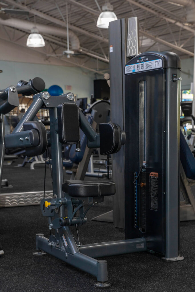 A seated rotary torso exercise machine in a gym, featuring padded arm and back supports, an adjustable weight stack, and various gym equipment in the background under bright overhead lights.