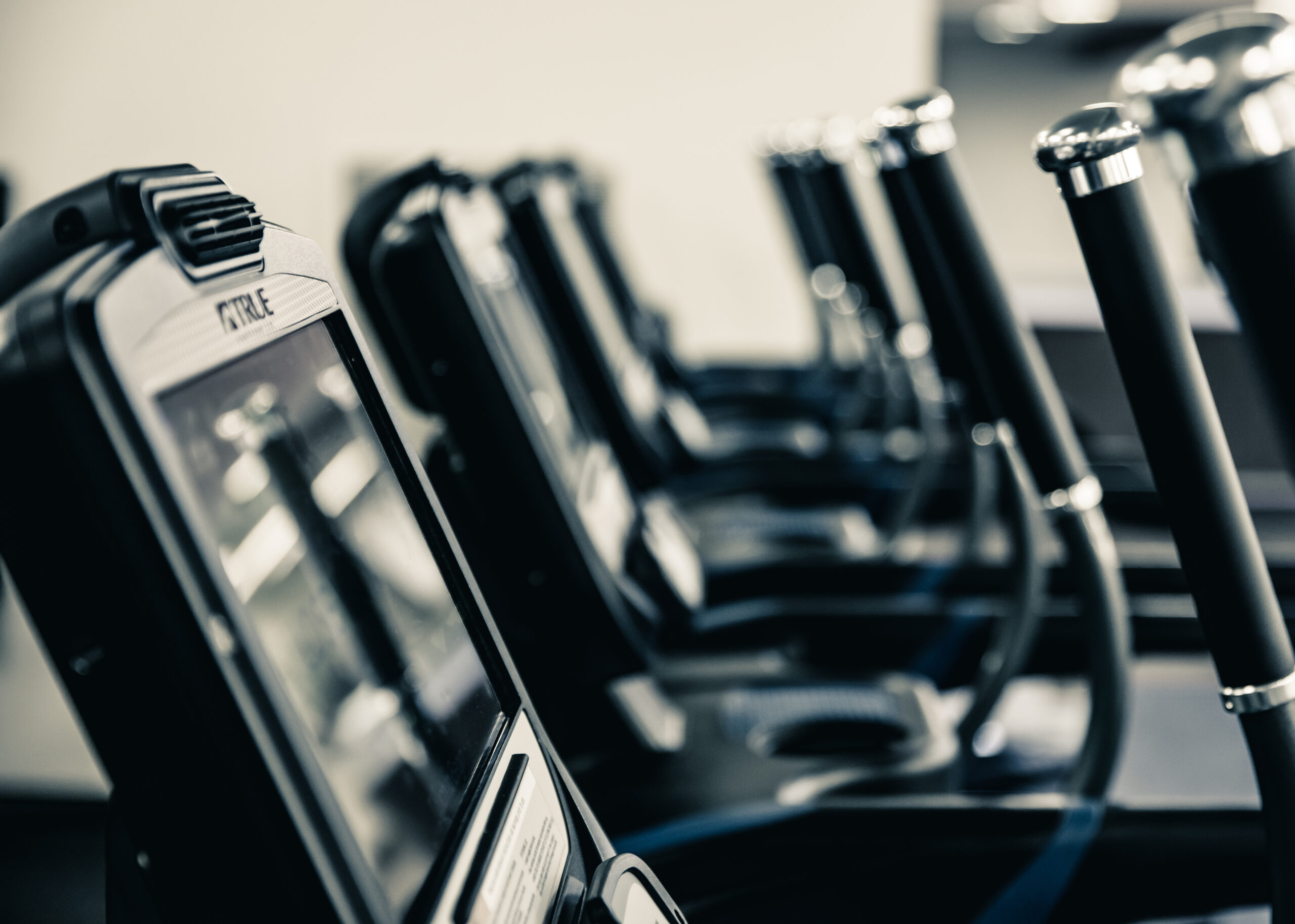 Close-up view of a row of exercise bikes or cardio machines in a gym, showing their screens and handlebars, with a shallow depth of field and a blurred background.