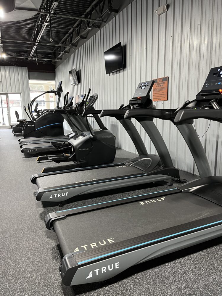 Row of treadmills and elliptical machines in a gym with gray walls, carpeted floor, and wall-mounted TVs. The room is empty and well-lit, with gym equipment neatly arranged side by side.