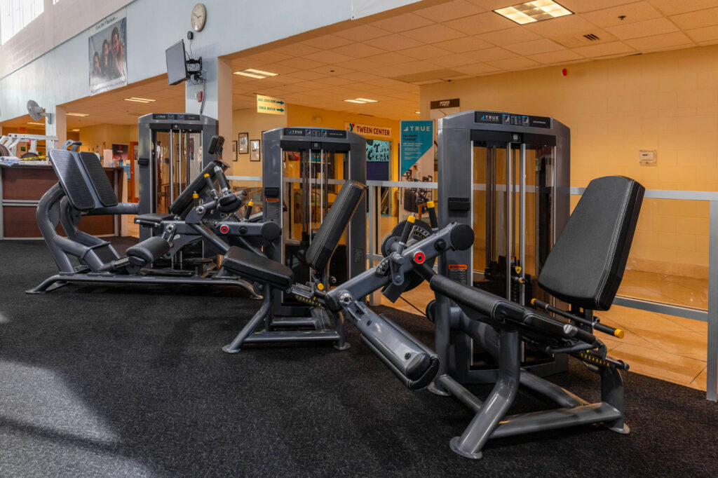 Three black leg press machines are lined up on a black carpeted floor in a gym with beige walls and overhead fluorescent lighting. Other gym equipment and a reception area are visible in the background.