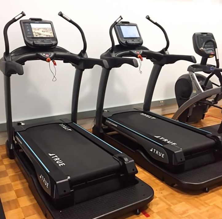 Two black treadmills with digital screens are positioned side by side on a wooden floor in a gym. An elliptical machine is partially visible to the right.