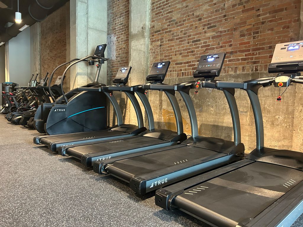 A row of treadmills and an elliptical machine in a modern gym with exposed brick walls and concrete pillars, all exercise equipment appears unused.