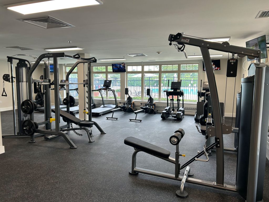 A modern gym with various fitness equipment including weight machines, benches, treadmills, and elliptical machines, with large windows and TVs mounted on the walls.