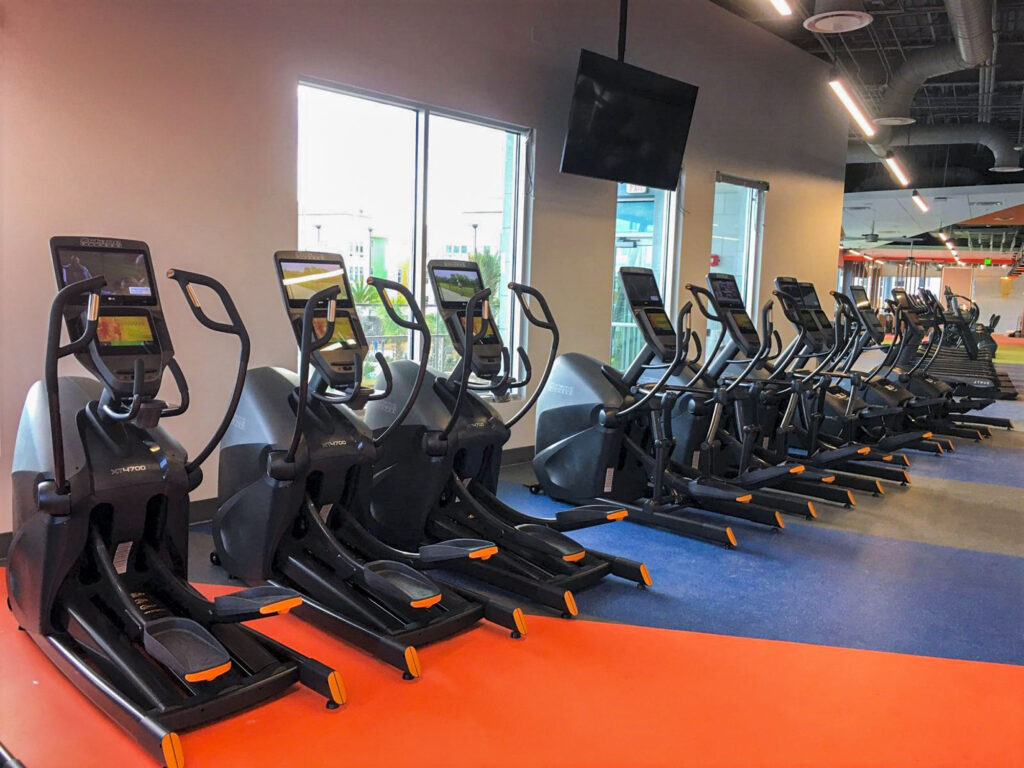 A row of elliptical machines is lined up in a modern gym with large windows, blue and orange flooring, and a TV mounted on the ceiling. The area is brightly lit and organized.