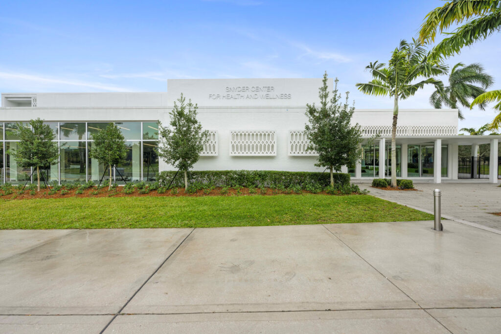 Modern white building labeled Sander Center for Health and Wellness, surrounded by palm trees and greenery, with a clear blue sky and clean, wet pavement in front.