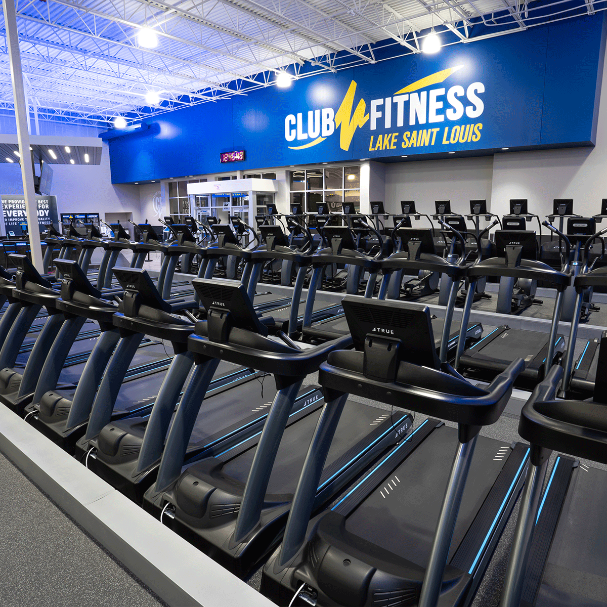 Rows of empty treadmills are lined up in a brightly lit gym with a large blue sign reading CLUB FITNESS LAKE SAINT LOUIS on the wall in the background.