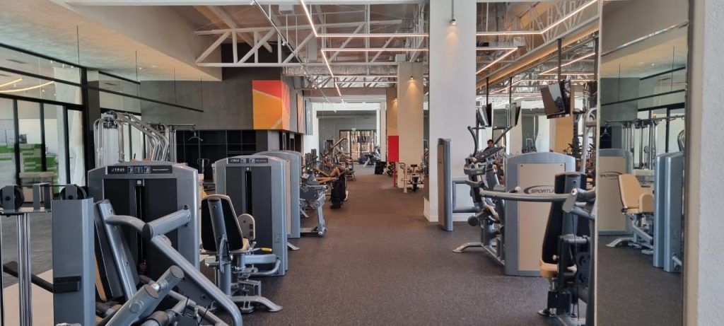 A spacious, modern gym with various strength-training and cardio machines arranged neatly. Large mirrors, high ceilings, and abstract art on the walls create a clean, open, and well-lit environment.