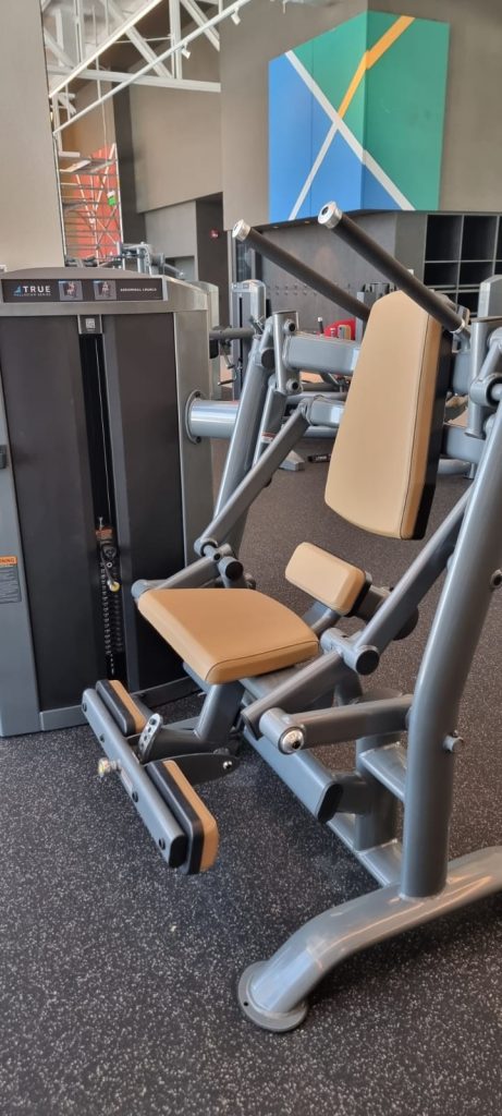 A shoulder press machine with tan padding and gray metal frame is positioned on a speckled black gym floor. A colorful geometric wall art and other gym equipment are visible in the background.
