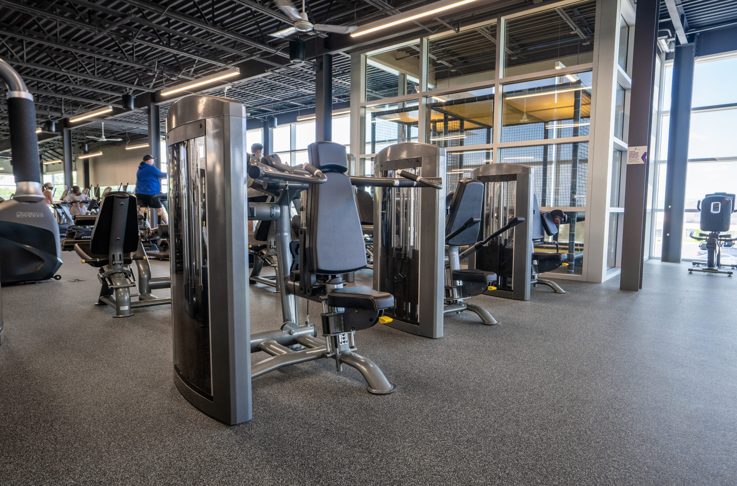 A modern gym interior with various strength training machines, including chest press and leg machines, arranged on a gray rubber floor. Large windows let in natural light, and a few people are seen working out in the background.