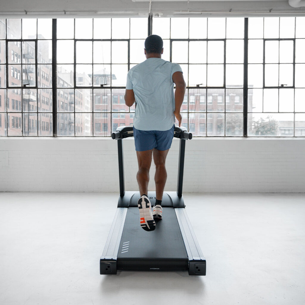A person in a light blue shirt and shorts runs on a treadmill in a bright, spacious room with large windows overlooking city buildings. The scene is minimalist, with natural light filling the space.