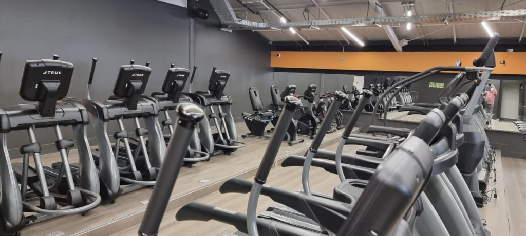 Rows of elliptical machines and exercise bikes are lined up in a modern gym with wooden flooring, gray walls, an orange accent stripe, and a mirrored wall reflecting the equipment and a person.