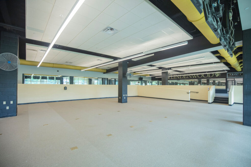 A spacious, modern indoor area with light-colored flooring, black ceiling beams, large windows, and yellow ventilation ducts. The walls are light with some black accents, and Lindenwood is visible on a sign in the background.