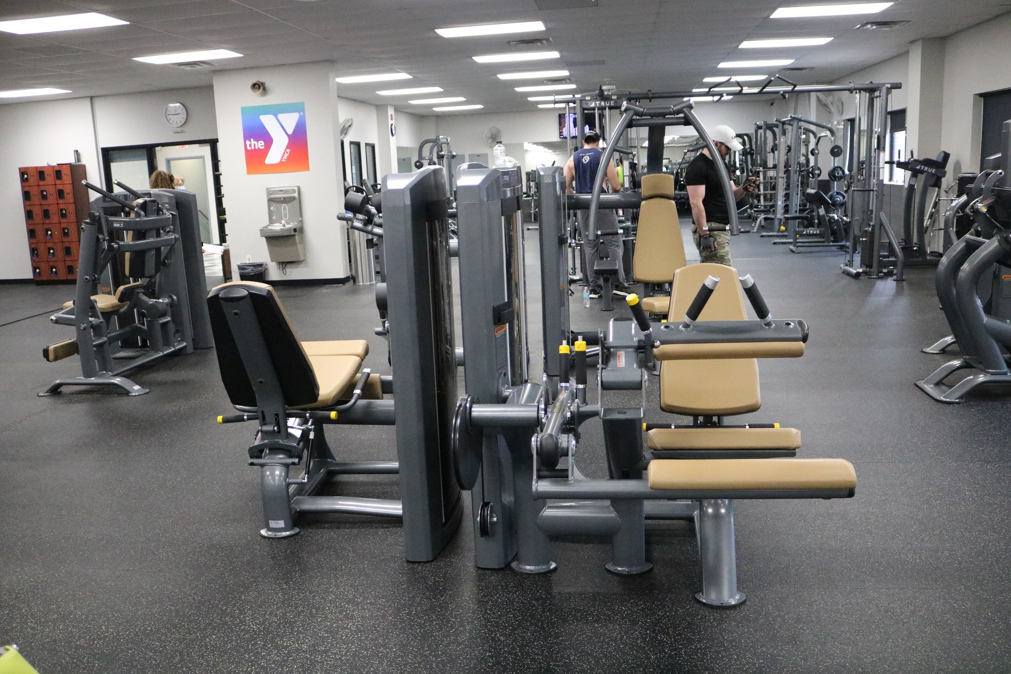 A gym with various exercise machines and equipment arranged in rows, tan and gray in color. The room is well-lit, with a Y logo on the wall and a few people working out in the background.