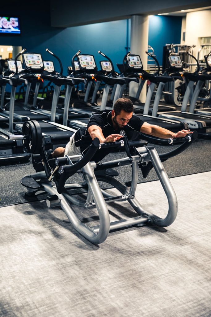 A man in athletic wear uses a chest-supported machine at the gym, extending his arms forward while lifting weights. Several treadmills are lined up in the background.