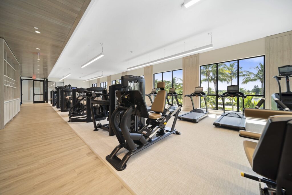 Modern gym with cardio and strength machines, large windows letting in natural light, and views of palm trees outside. The room has light wood flooring and a clean, spacious layout.