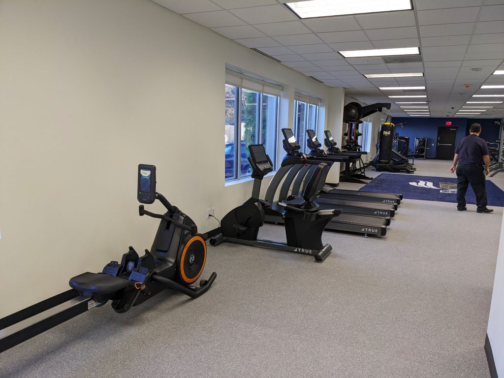 A gym room with exercise machines including a rowing machine, stationary bikes, treadmills, and ellipticals. Large windows line one wall, and a person is walking away in the background.