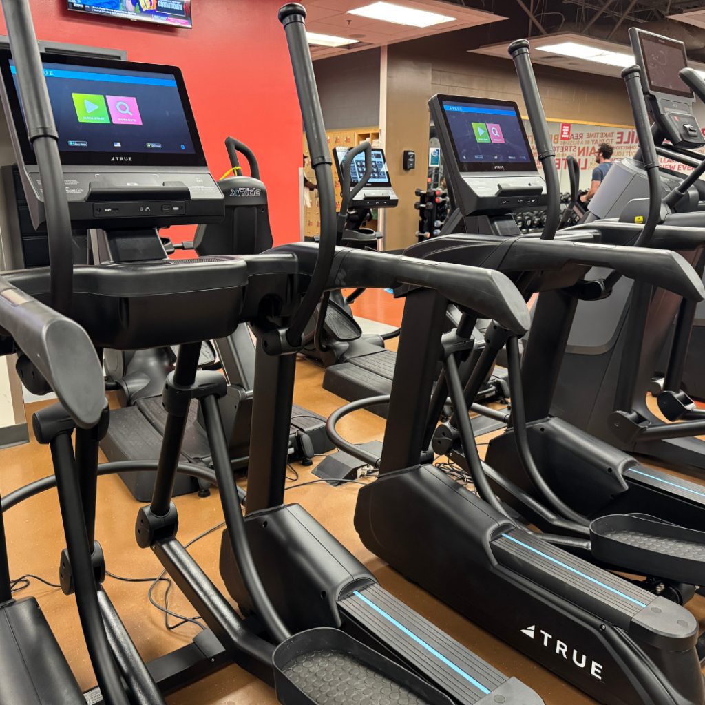 A row of elliptical machines with digital screens is lined up in a gym. Some screens display colorful icons. The gym has brown flooring and bright lighting. Other fitness equipment is visible in the background.
