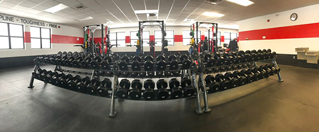 A gym with rows of dumbbells arranged on racks in front of power racks. The room has red, white, and black walls, a clock, windows, and motivational words displayed above the windows.