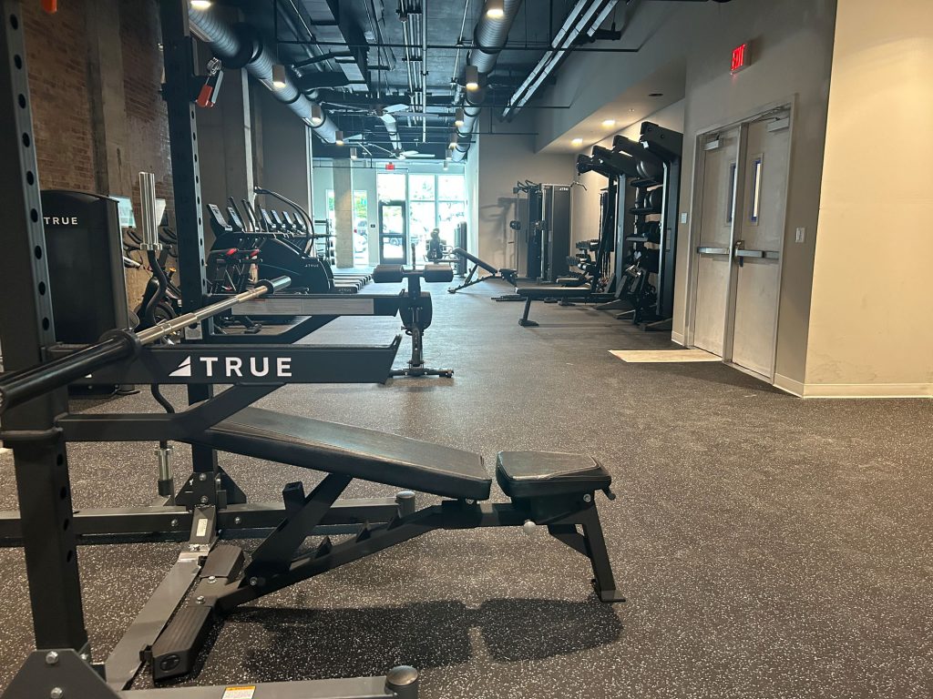 A modern gym with various exercise equipment, including benches, weight racks, and cardio machines, arranged neatly in a spacious room with rubber flooring and exposed ceilings.