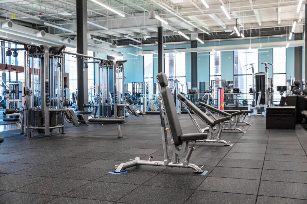 A spacious, modern gym with weight machines, benches, and exercise equipment arranged on rubber flooring under bright overhead lighting and large windows.