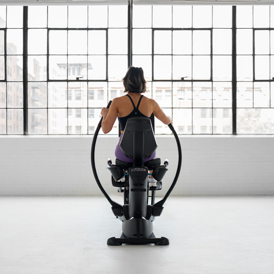 A woman in athletic wear uses a rowing machine in a bright, spacious room with large industrial-style windows and a cityscape view.