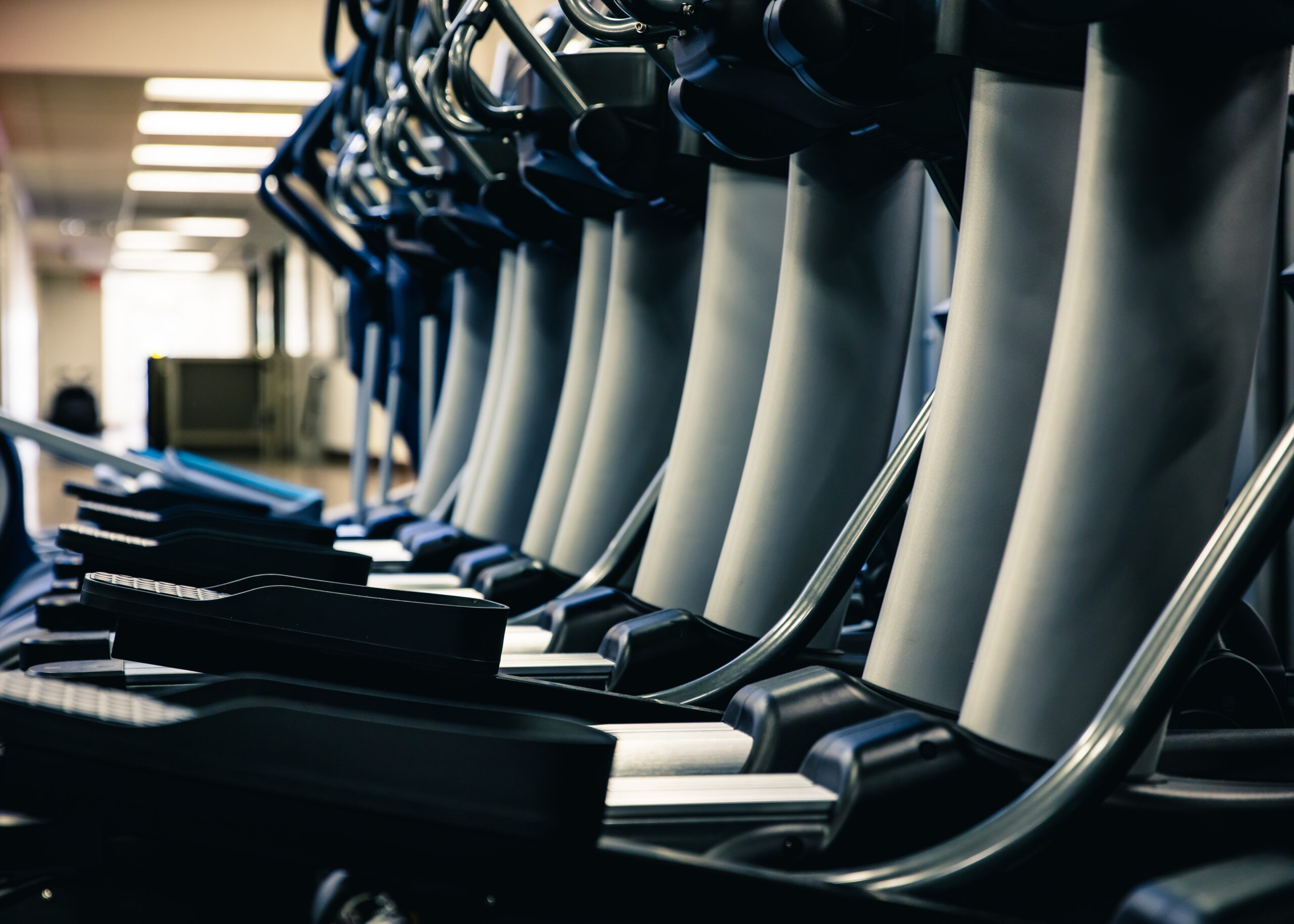 A row of elliptical exercise machines lined up in a gym, with a blurred background showing bright overhead lights and gym equipment.
