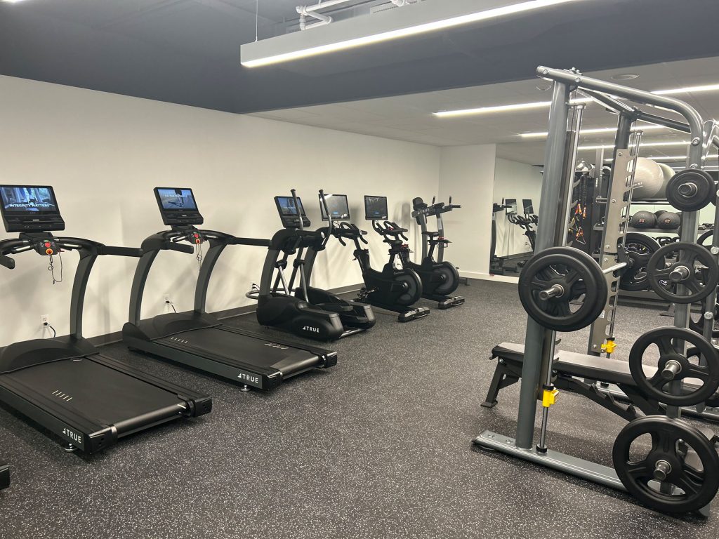 A modern gym with treadmills, stationary bikes, and weightlifting equipment. Each treadmill has a screen, and the room features mirrors on one wall, creating a clean, organized workout space.