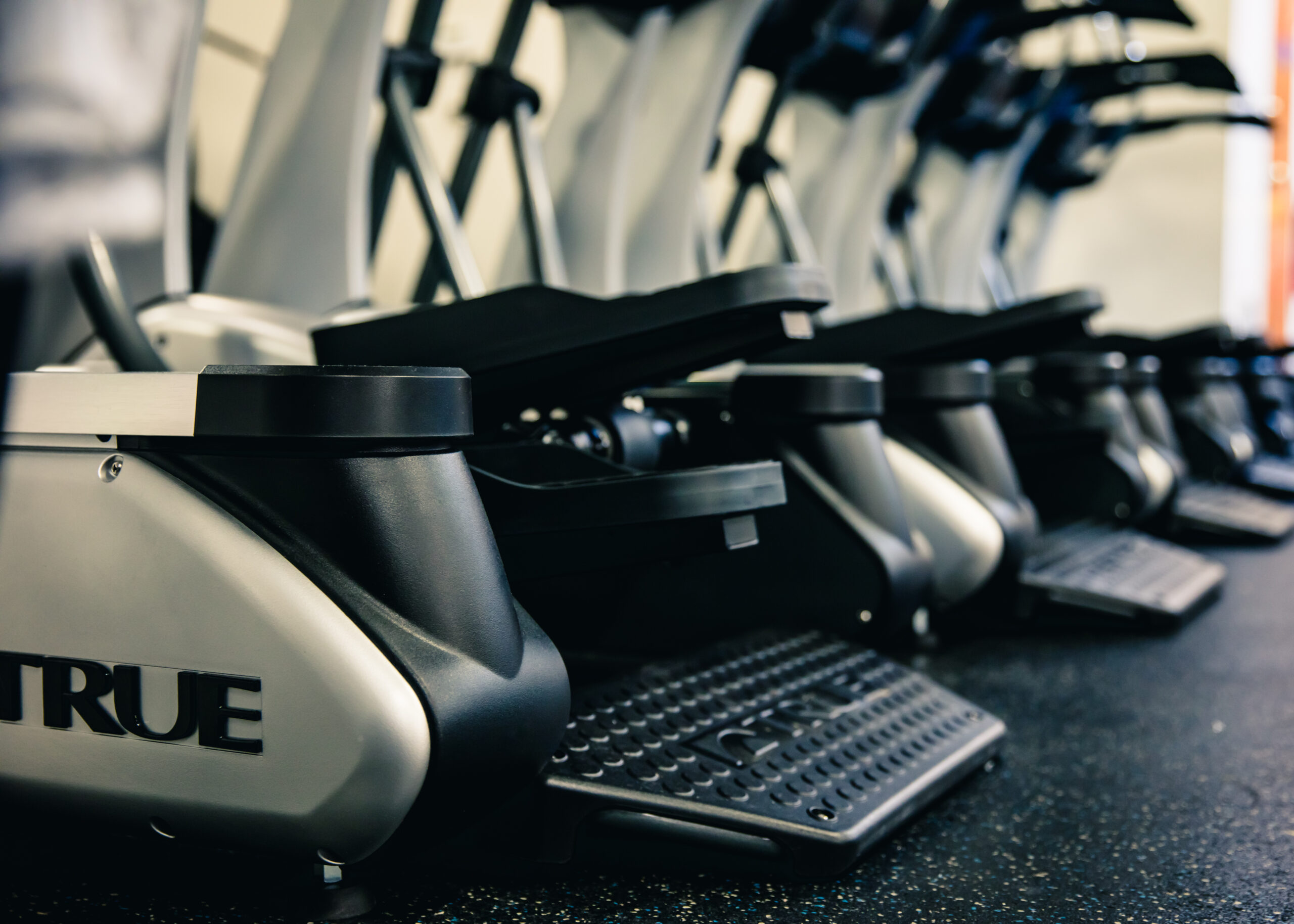Close-up of several elliptical machines lined up in a gym, focusing on the pedals and lower frame. The floor is covered in rubber gym flooring. The word TRUE is visible on the side of one machine.