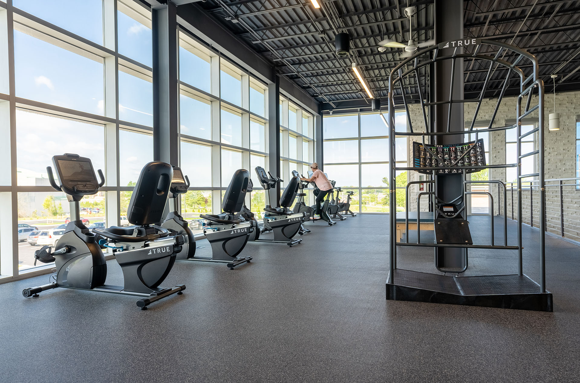 A spacious, modern gym with large windows, several recumbent exercise bikes, and a person using one bike. Natural light fills the space, highlighting the clean, open workout area.