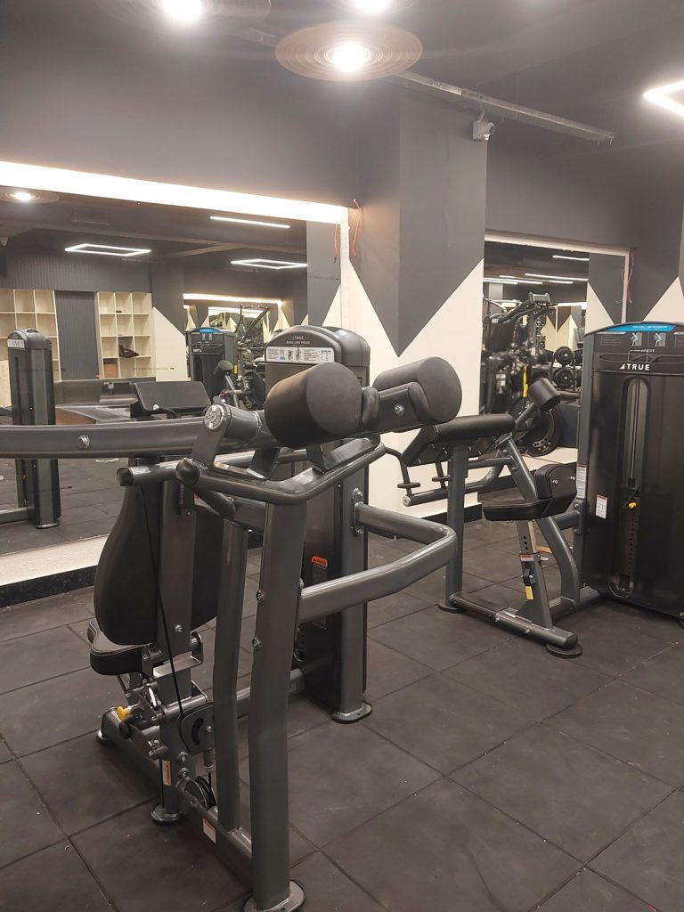A gym with various exercise machines on black flooring, mirrored walls, and shelves in the background. The lighting is bright, and the area appears clean and organized, with no people present.