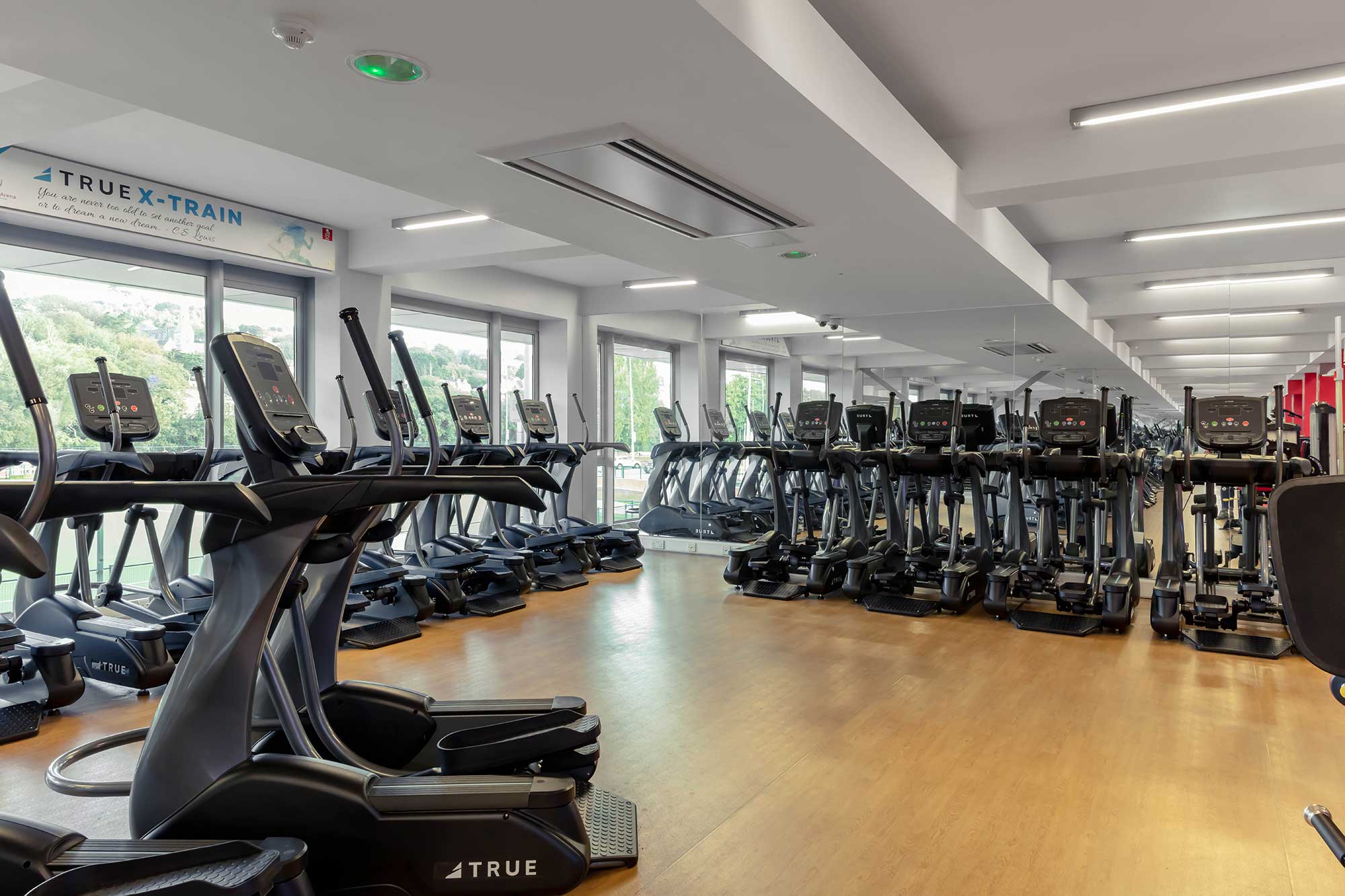 A spacious, modern gym with rows of treadmills, ellipticals, and exercise bikes on a wooden floor, surrounded by large windows letting in natural light.