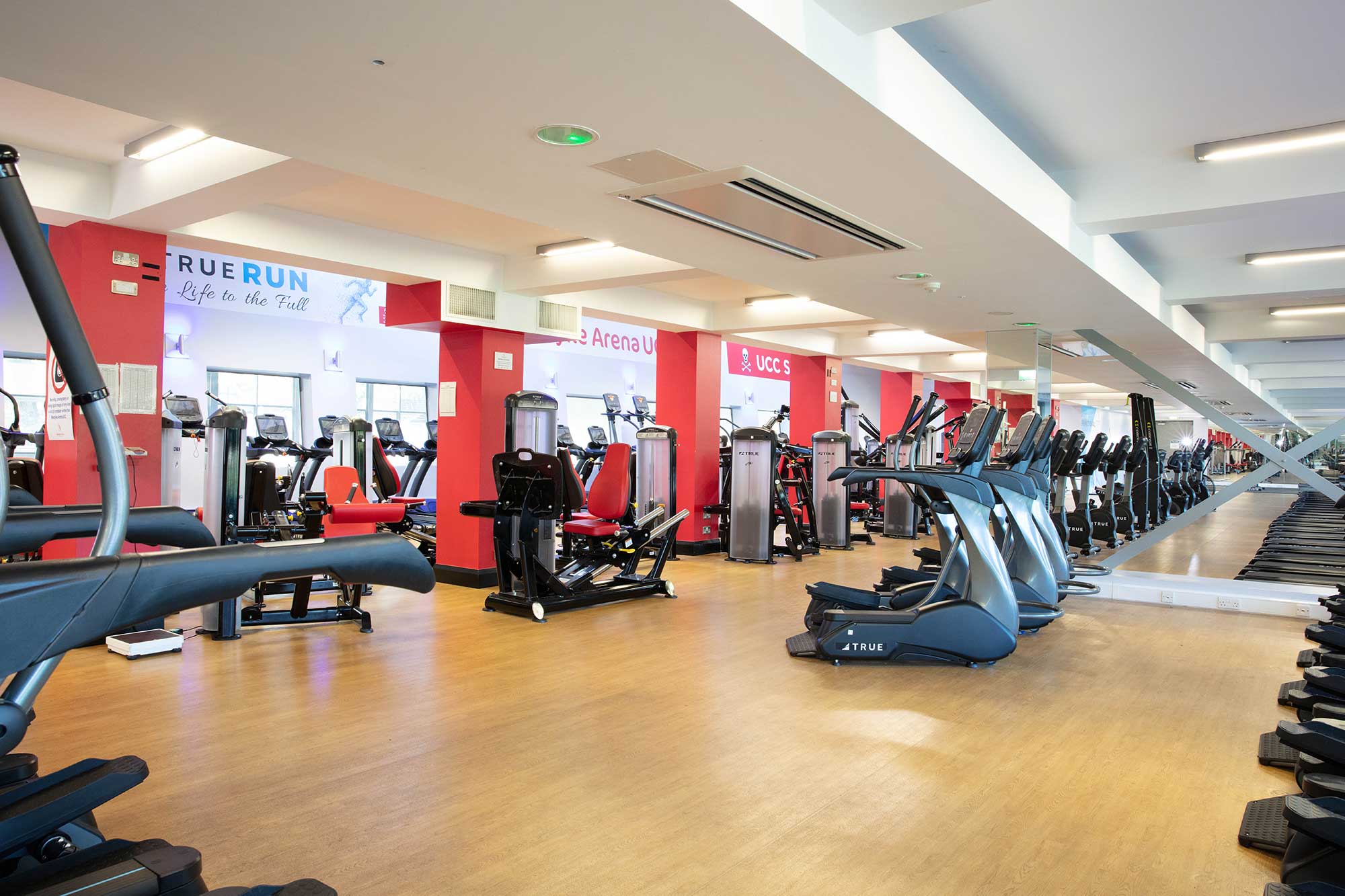 A spacious gym with wooden floors, red and white walls, and large mirrors. Rows of treadmills and various fitness machines are neatly arranged, with bright lighting and motivational signs on the walls.