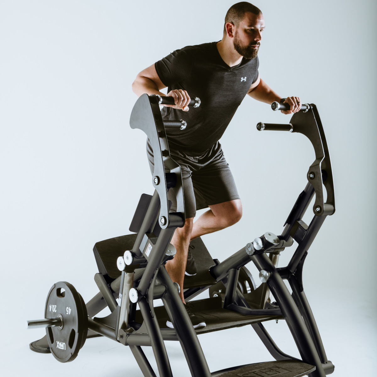 A man wearing a black athletic outfit uses a leg press machine in a gym, gripping the handles and positioning his feet on the platform as he prepares to exercise.