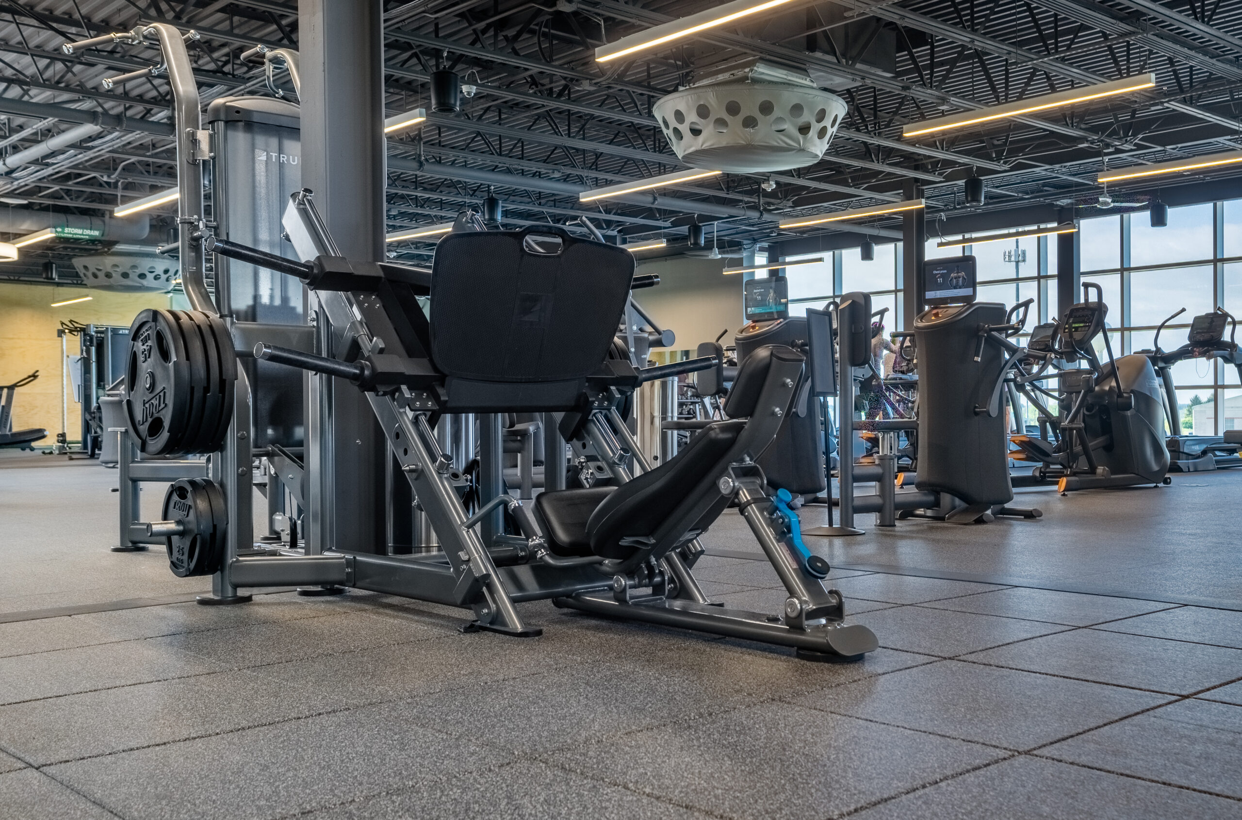 A modern gym with various exercise machines, including a leg press in the foreground, ellipticals, and treadmills in the background. The space has large windows and overhead lighting.
