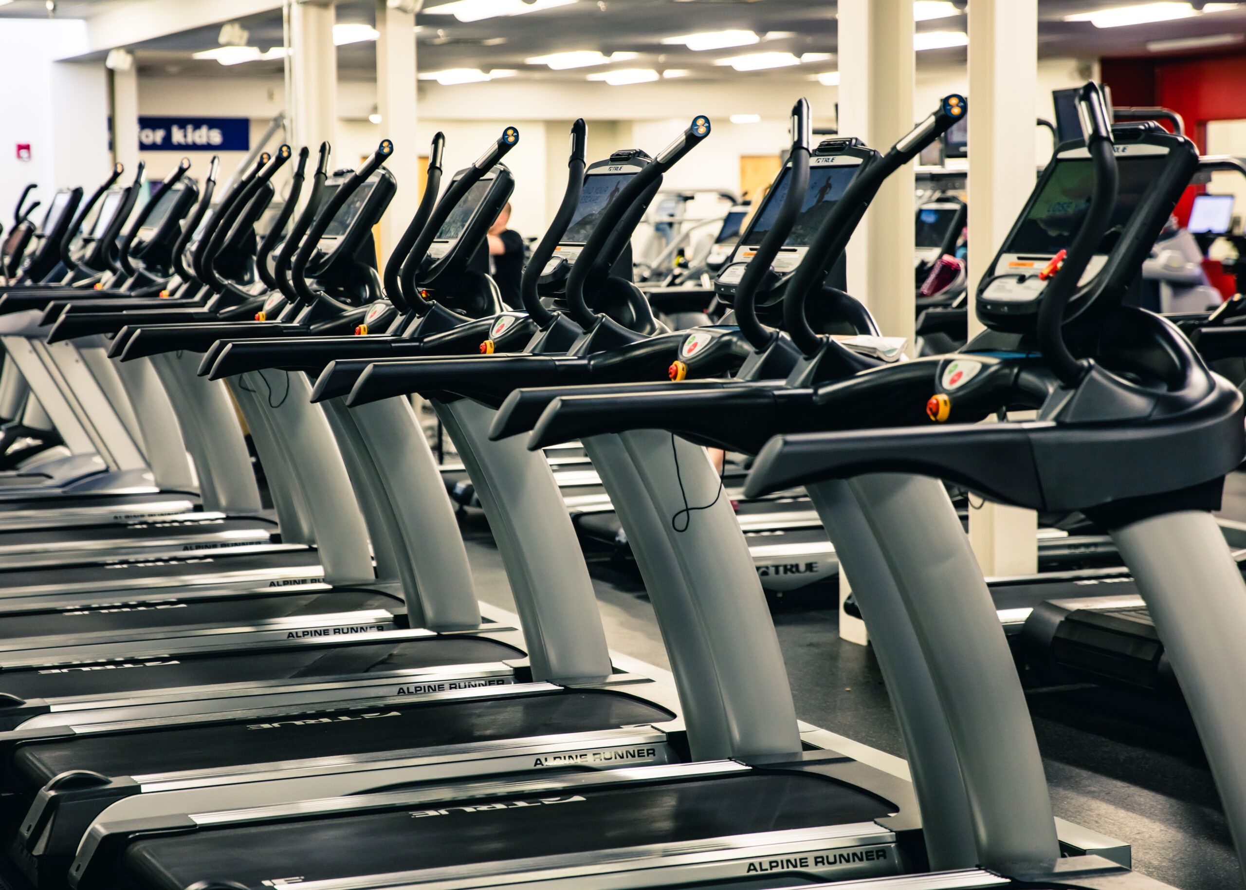 A row of treadmills lined up in a brightly lit gym, with exercise equipment and people visible in the background.