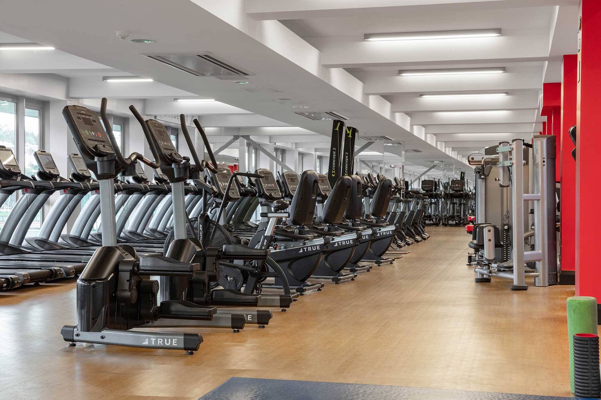 A spacious, well-lit gym with rows of treadmills, elliptical machines, stationary bikes, and strength training equipment on wooden flooring. The area appears clean and organized, with large windows along one side.