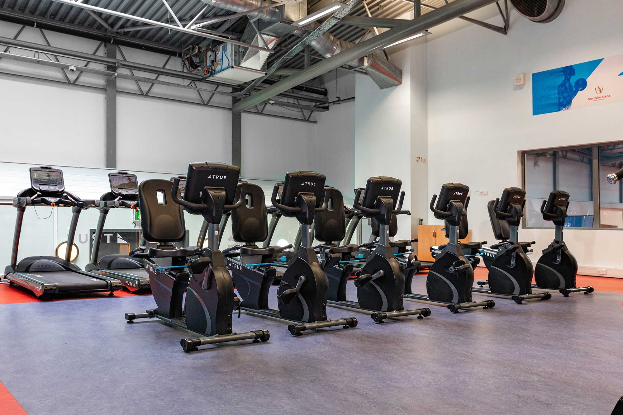 A row of six stationary exercise bikes and two treadmills are lined up in a spacious, modern gym with high ceilings, exposed ducts, and bright lighting.