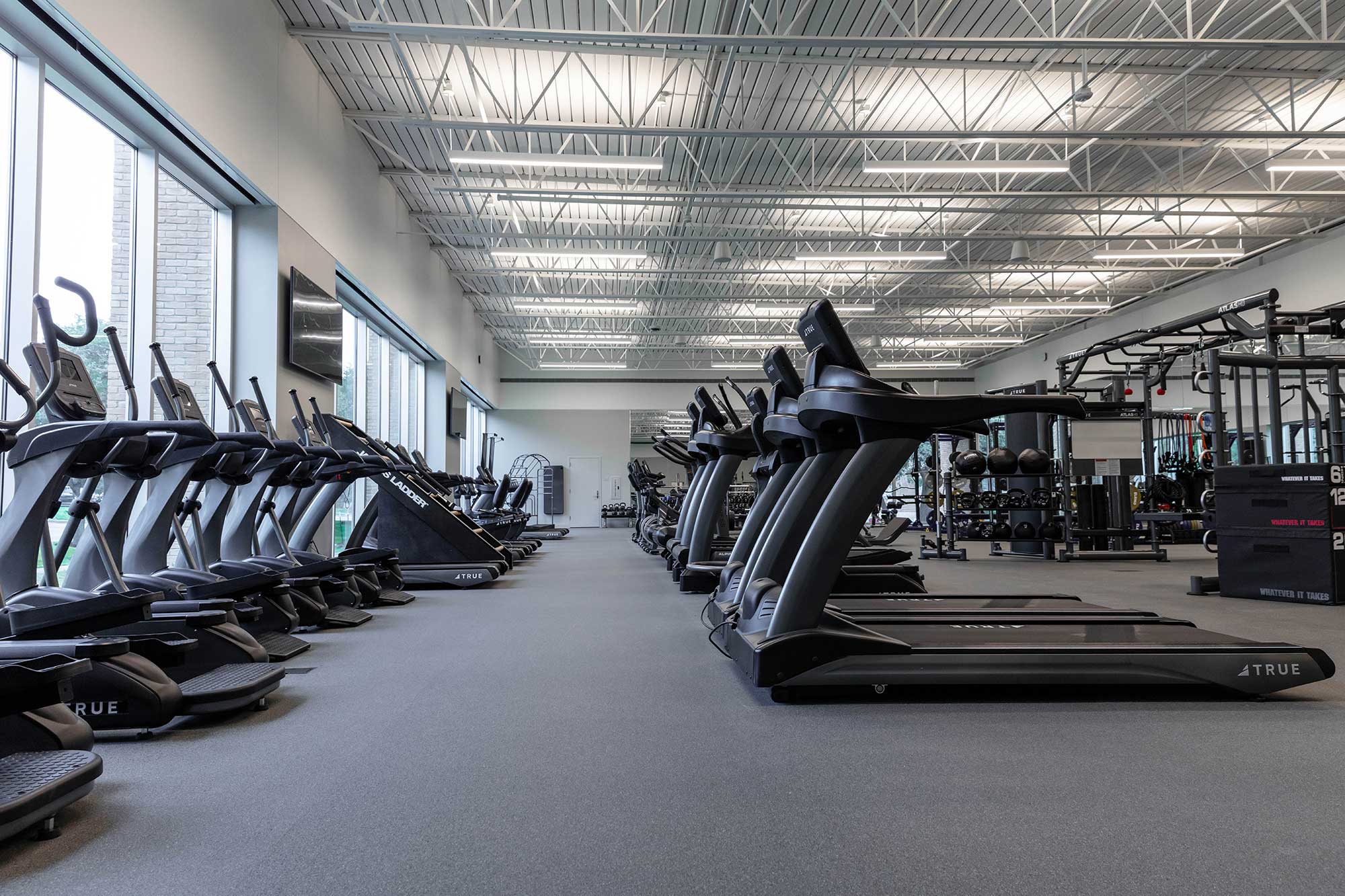 A spacious, modern gym with large windows, filled with rows of treadmills, ellipticals, and other cardio machines on one side, and strength training equipment on the other. The gym has high ceilings and bright lighting.