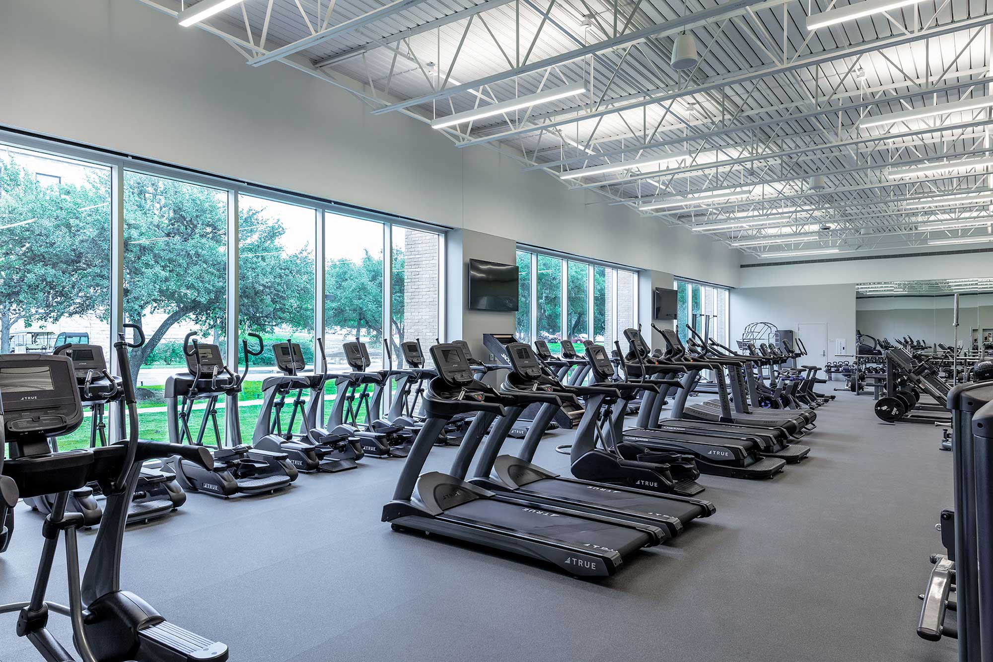 A bright, spacious gym with large windows, filled with rows of treadmills and elliptical machines, all facing the windows. Trees and greenery are visible outside. The gym has high ceilings and a clean, modern look.