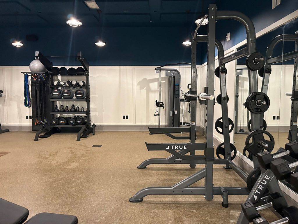 A clean gym with weight racks, dumbbells, exercise balls, resistance bands, and strength training equipment against white walls and a blue ceiling. The space is well-lit and organized.