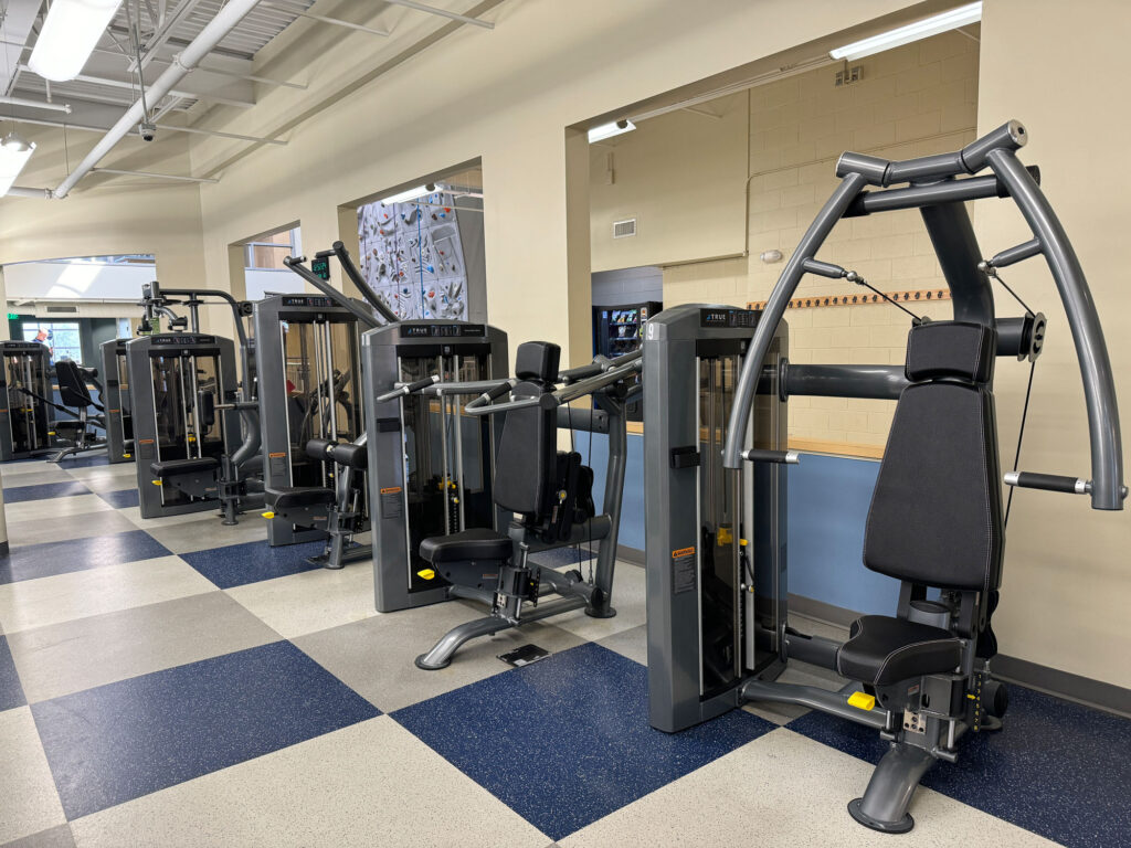 A row of strength training machines in a gym, featuring padded seats and metal frames, set on a checkered blue and gray floor with mirrors and climbing wall visible in the background.