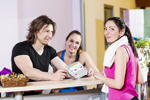 Three people at a gym reception desk talk and smile together. A man and a woman behind the counter hold a schedule, while a woman with a towel around her neck stands on the other side, facing them.