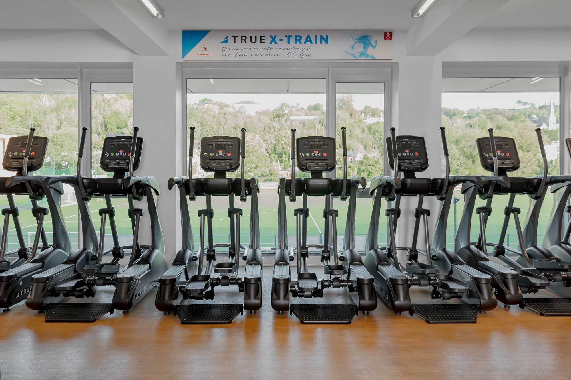 A row of six elliptical machines in a bright gym with large windows overlooking greenery; a banner above reads TRUE X-TRAIN.