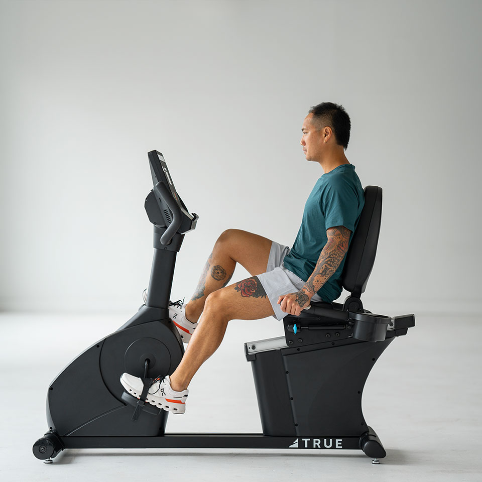 A man in a teal shirt and white shorts uses a recumbent exercise bike in a bright, minimalist room. He is seated upright, facing forward, with one tattooed arm visible. The bike is labeled TRUE.