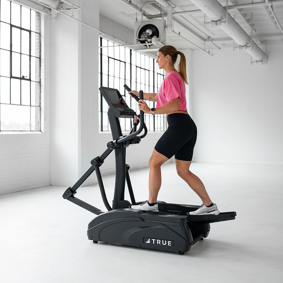 A woman in a pink shirt and black shorts exercises on a TRUE elliptical machine in a bright, spacious gym with large windows and white walls.