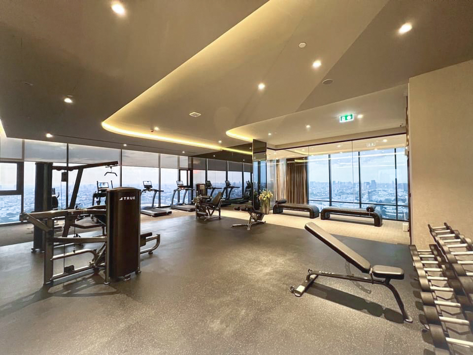 A modern gym with large windows offering a city view, featuring treadmills, ellipticals, weight machines, benches, and a rack of dumbbells in a spacious, well-lit area.