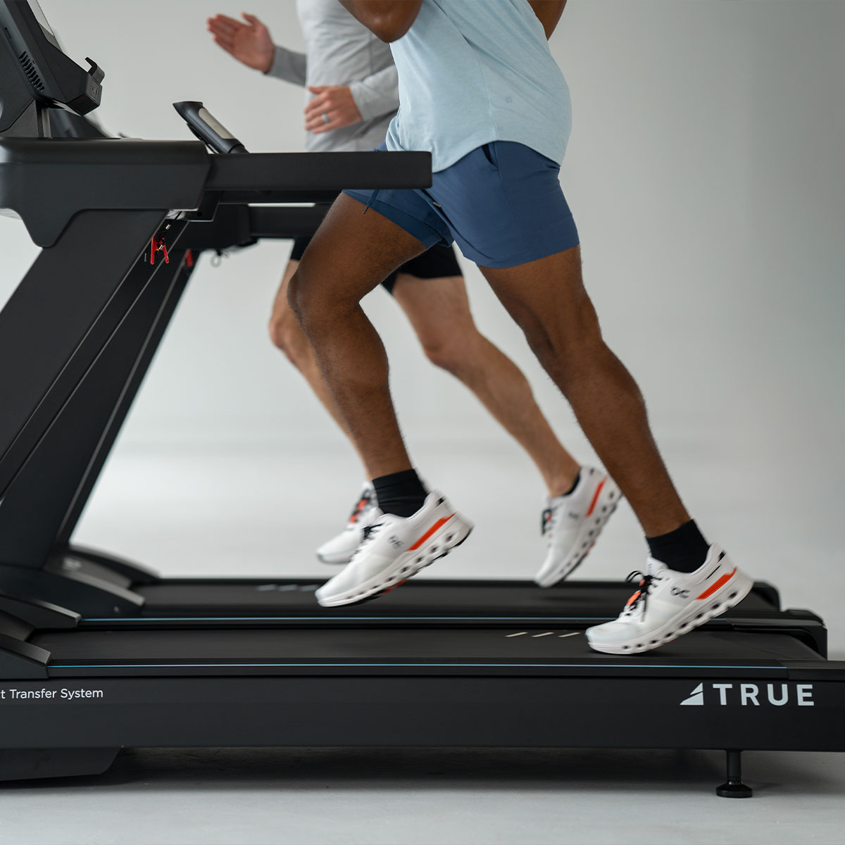 Two people run side by side on treadmills in a gym, wearing athletic shorts, shirts, and white running shoes. The focus is on their legs and the treadmill brand “TRUE” is visible.